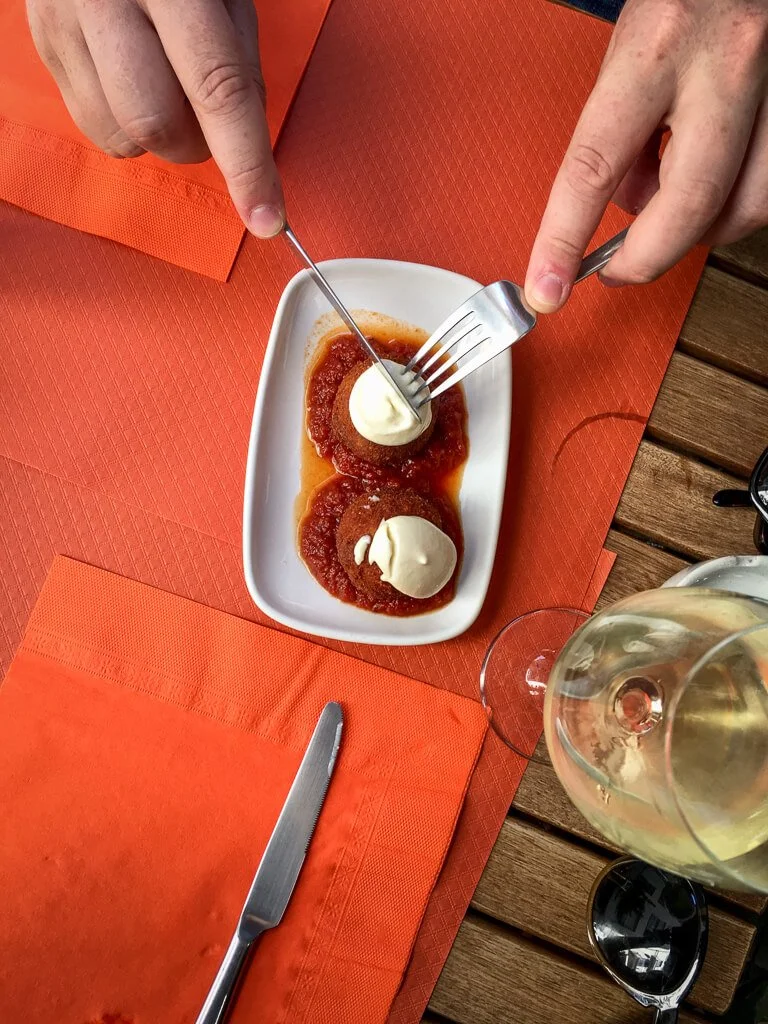 A rectangular white plate holding two round, golden-brown croquettes served on a bed of tomato sauce, each topped with a dollop of white aioli. A person is using a knife and fork to cut one open, with a glass of white wine nearby on an orange tablecloth.