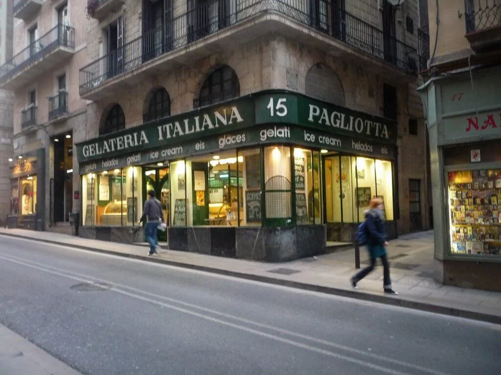 A street view of an old-fashioned corner gelato shop with a green awning reading “Gelateria Italiana” and “Pagliotta.” The sidewalk has a couple of people walking by, and the shop’s warm lights glow from inside.