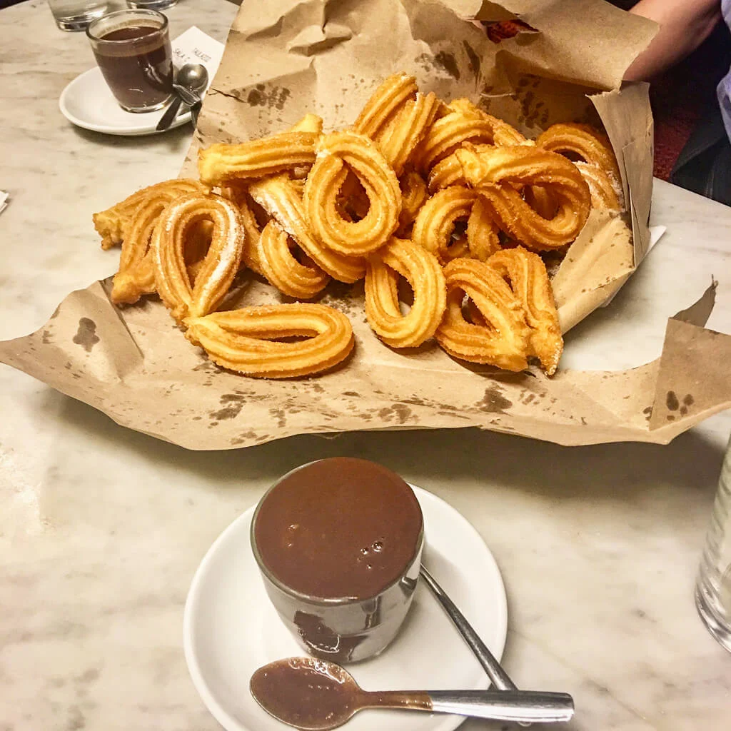 A pile of freshly fried churros arranged on brown parchment paper, placed on a marble table. In front of them is a small glass filled with thick hot chocolate on a white saucer with a spoon resting beside it, ready for dipping.