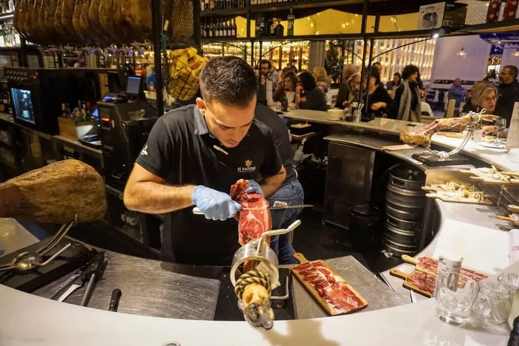 A man behind a bar counter carefully slicing a leg of cured ham mounted on a stand. He’s wearing gloves and focusing on thin, precise cuts. Plates with neatly arranged slices of ham are visible on the counter, and patrons sit in the background.