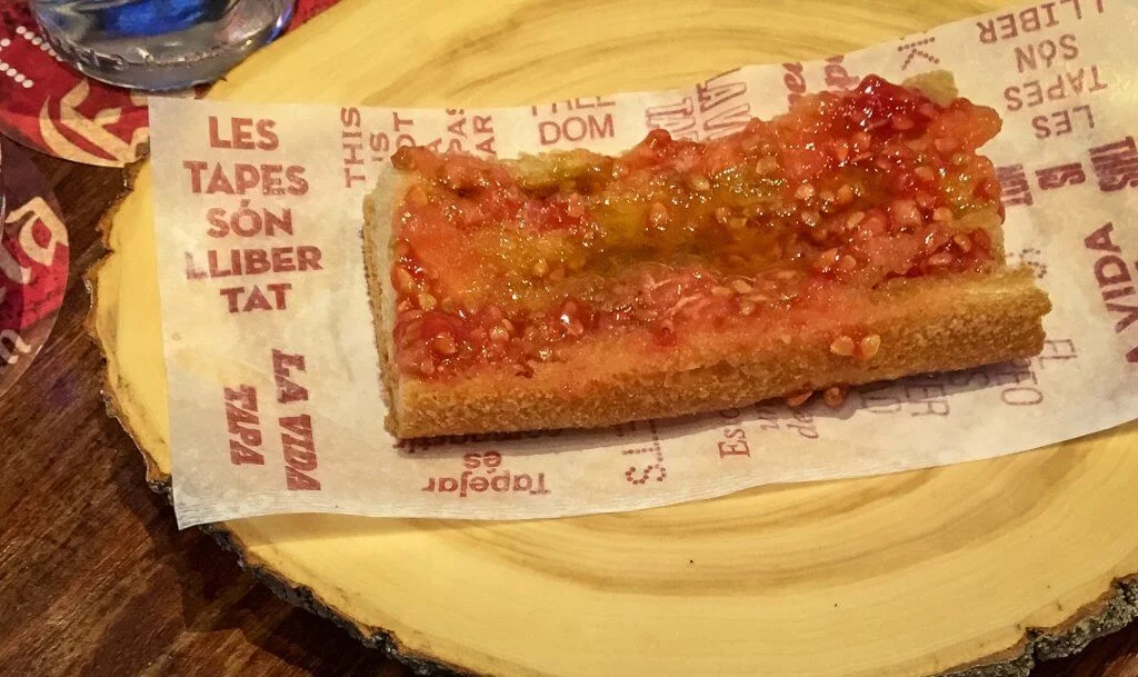 A slice of rustic bread topped with freshly grated ripe tomato, drizzled with olive oil. The bread is placed on branded paper atop a wooden serving board.