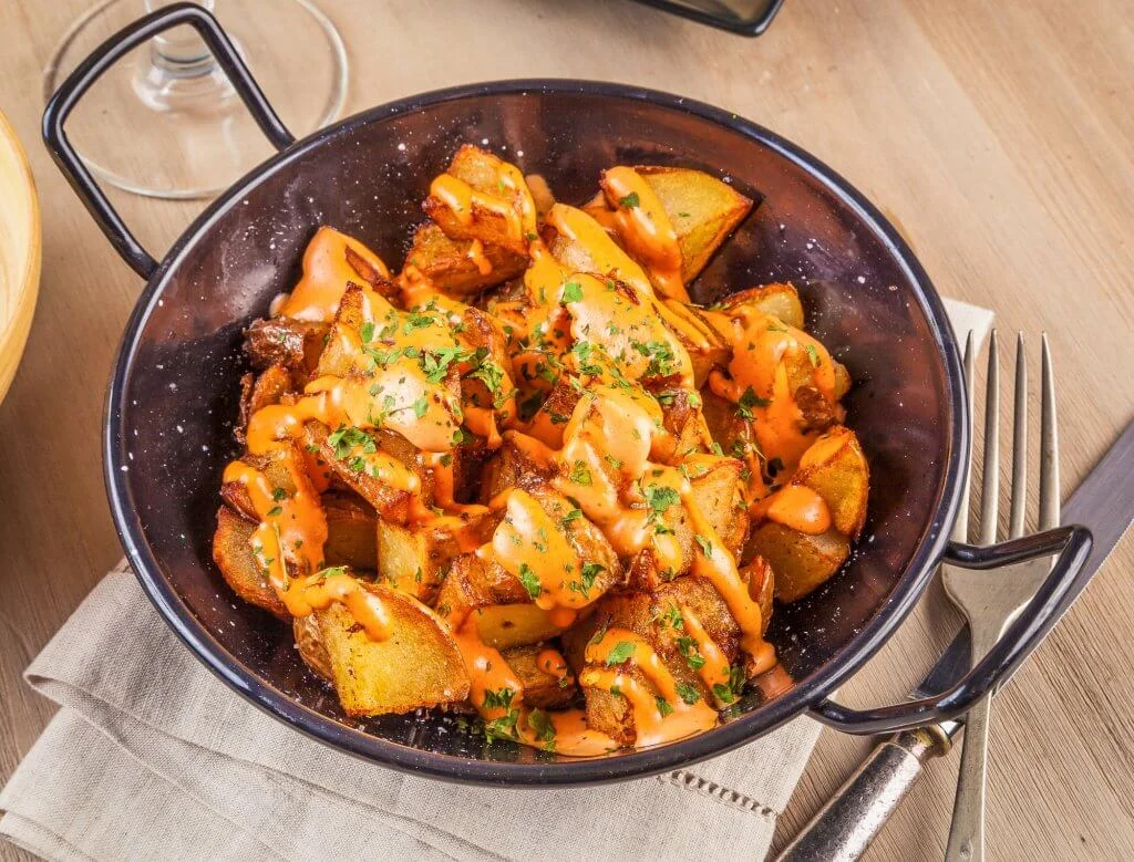 A black bowl filled with crispy golden-brown potato cubes, drizzled with an orange spicy aioli sauce and sprinkled with chopped parsley. The bowl is placed on a beige cloth napkin with a fork and knife beside it on a wooden table. A wine glass is partially visible in the background.
