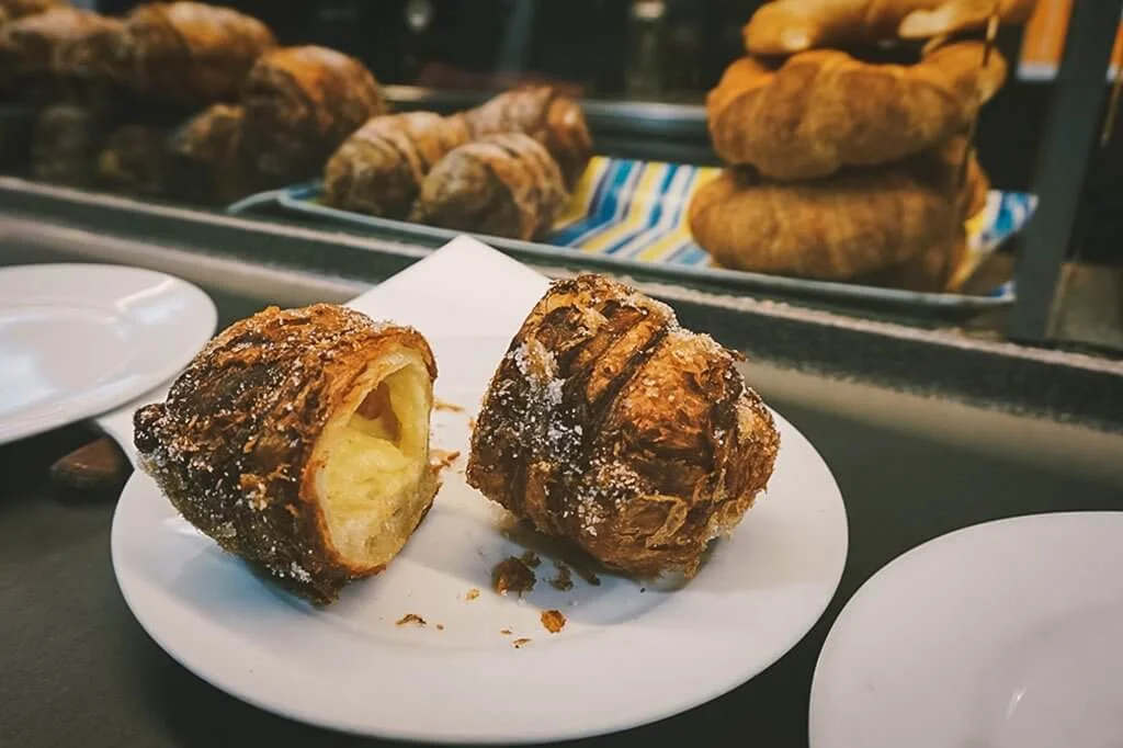 A white plate holding a sugar-dusted croissant cut in half, revealing a creamy custard filling inside. Flaky pastry crumbs are scattered on the plate. In the background, more croissants are stacked near a counter with a striped cloth underneath.