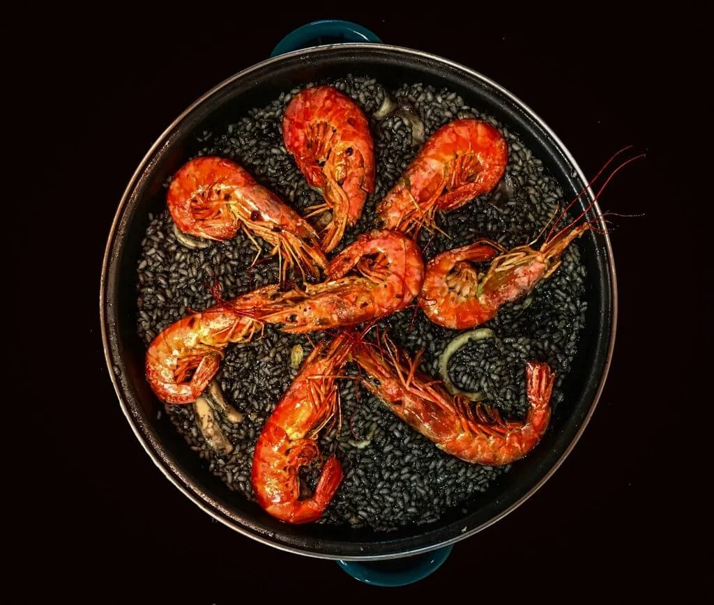 A black paella pan filled with black rice made with squid ink, topped with large bright orange prawns arranged in a circular pattern. The contrast between the dark rice and the vibrant prawns is striking, and a few pieces of squid are visible among the rice. The photo is taken from above against a black background.