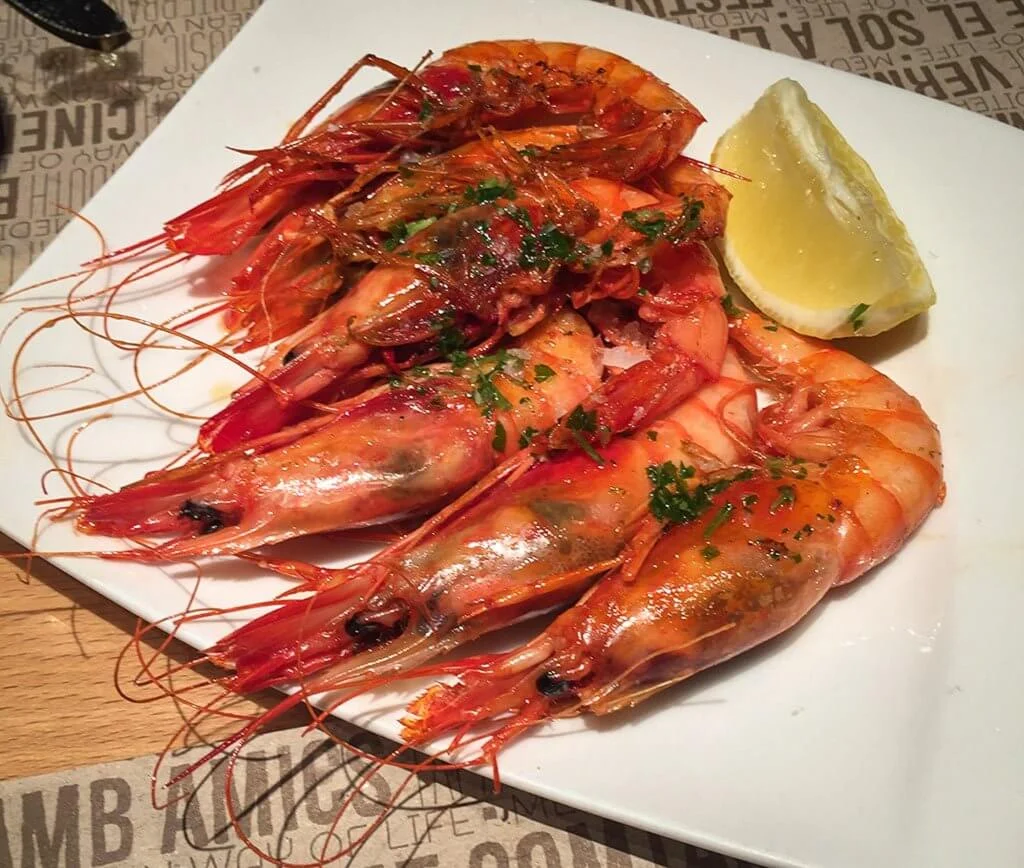A close-up of a white plate with several large grilled prawns garnished with chopped parsley and served with a wedge of lemon. The prawns glisten with oil and seasoning.