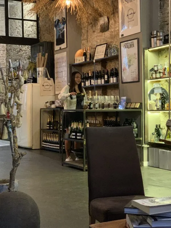 a woman pouring wine at a little shop, with various wine bottles on shelves next to her.