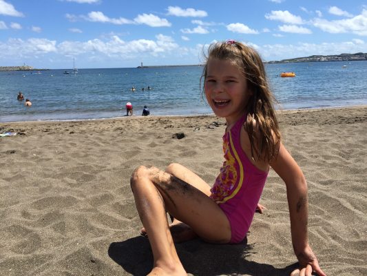 Girl sitting on a sandy beach in a pink swimsuit, laughing, with the ocean and blue sky behind her.