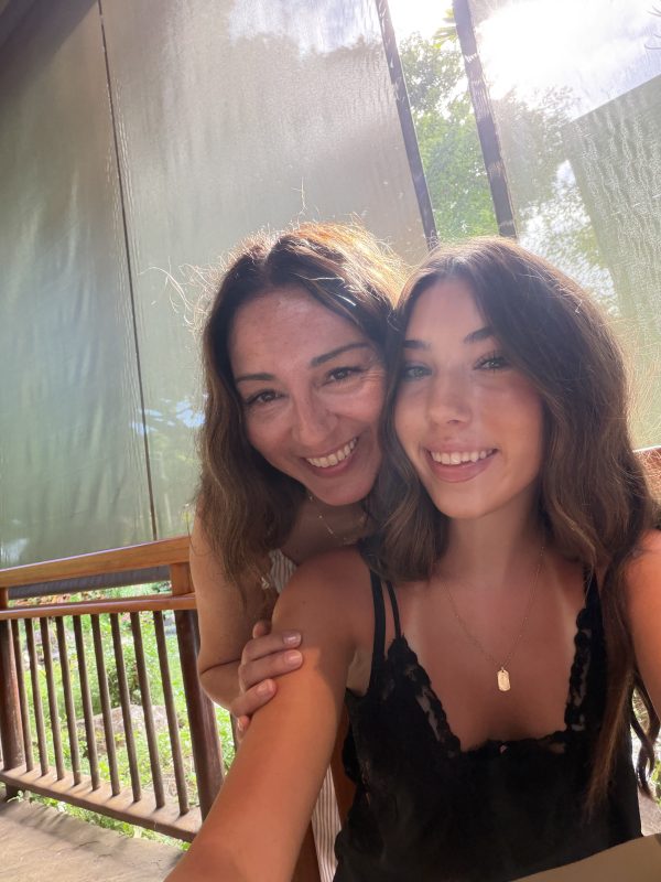 Two women smiling in a close selfie on a sunny outdoor porch in Kauai, green trees visible behind them.